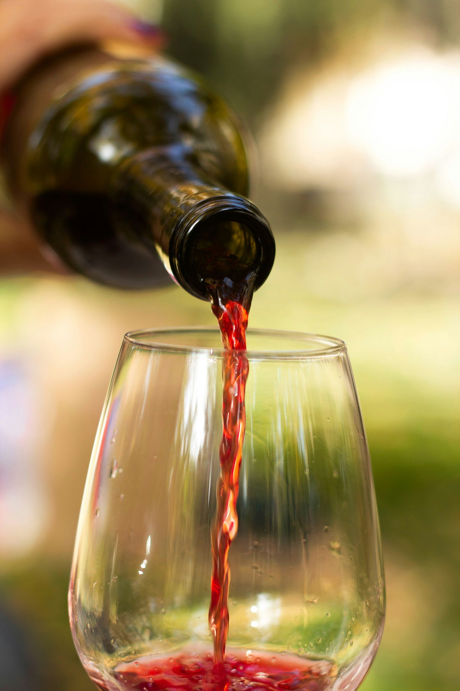 Rotwein fließt aus Flasche in ein Weinglas, sonniger Hintergrund.